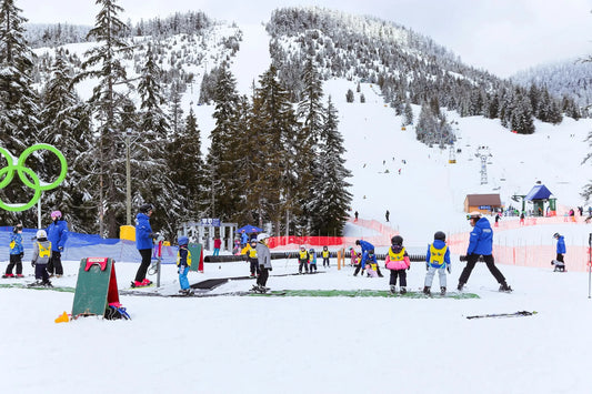 Children learning to ski on a snowy beginner slope with instructors, surrounded by pine trees and winter mountains.