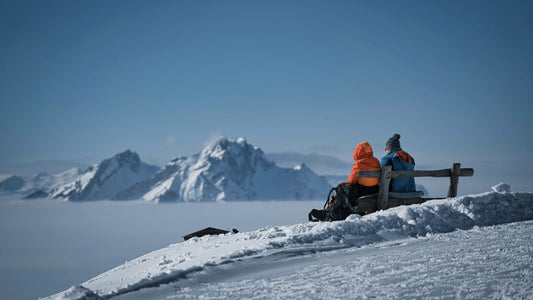 Two skiers sitting on a bench overlooking snowy mountains in a cold alpine environment, illustrating rest moments where foot warmth drops during skiing