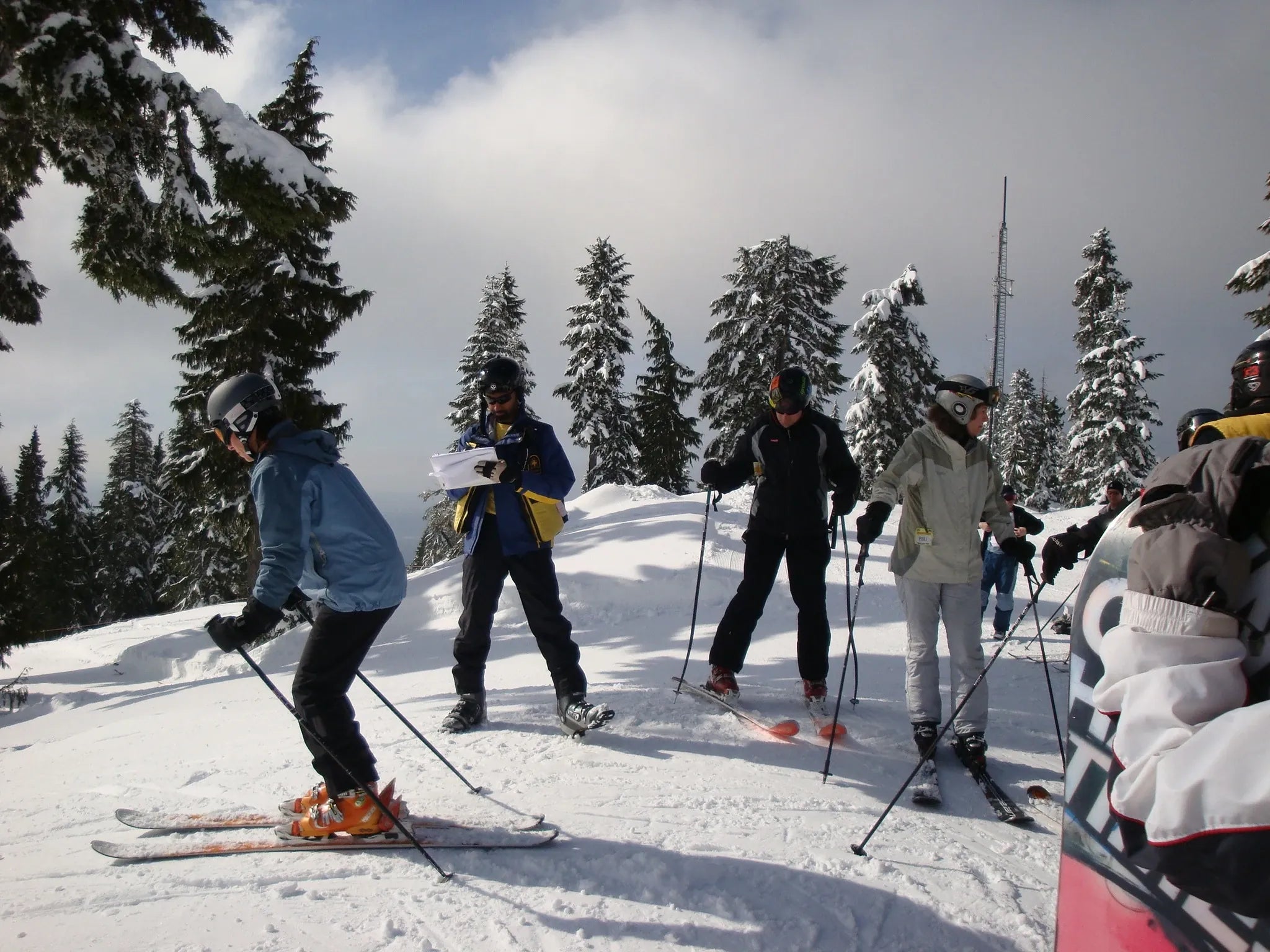 A group of skiers standing on a snowy mountain slope, preparing to ski. They are wearing snow gear and using ski poles, with snow-covered trees and a cloudy sky in the background.