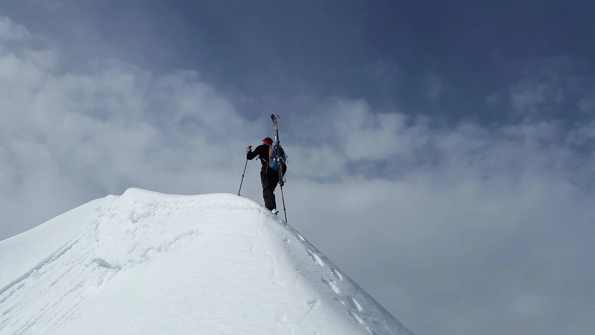 Winter hiker ascending a snowy ridge with skis on backpack, demonstrating cold-weather endurance and winter adventure performance.
