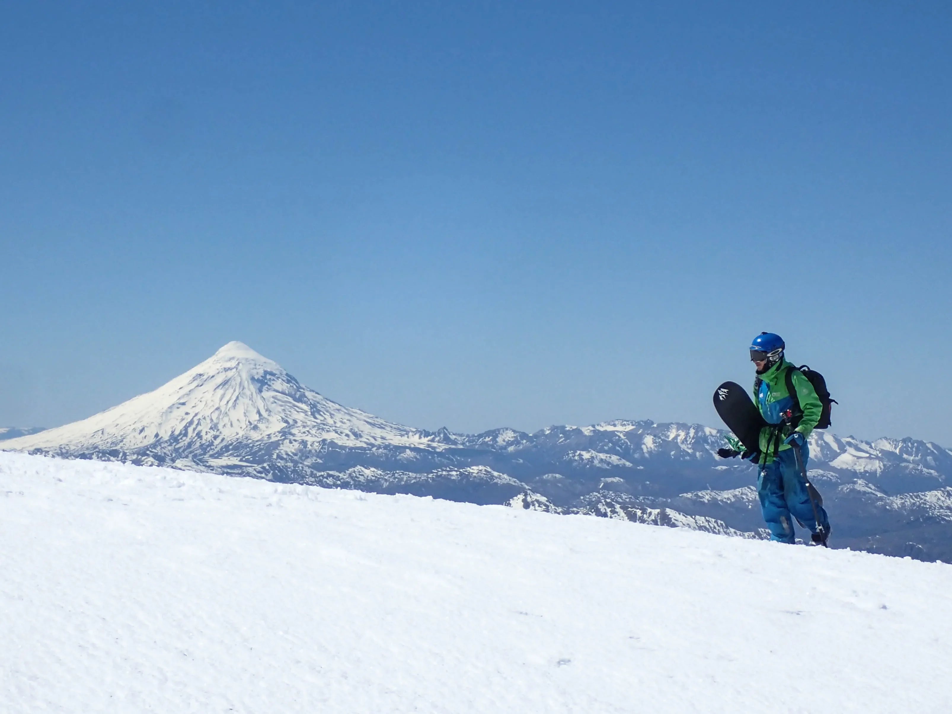 Snowboarder hiking up a snowy ridge with board in hand, framed by a distant alpine peak and clear blue sky—high-altitude winter adventure.